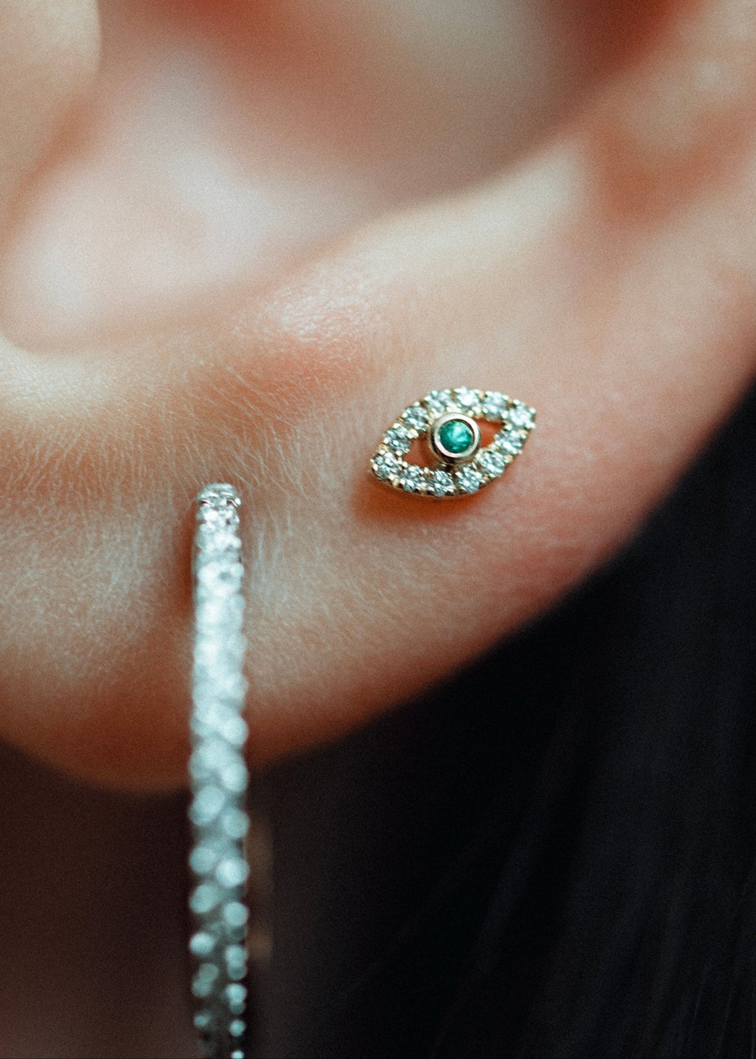 Extreme close-up of the Eirene Eye labret, a marquise of white diamonds around a green emerald, beside a pave diamond hoop.